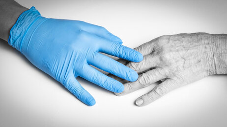 Bildet viser en hånd iført en plasthanske som berører hånden til et eldre menneske