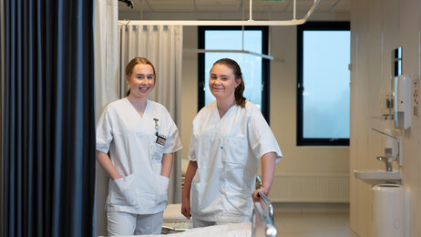 Bildet viser to sykepleiere på sengepost som ser mot kamera