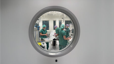Bildet viser en dør med et rundt vindu inn til en operasjonssal. Gjennom vinduet kan man se flere sykepleiere i arbeid.