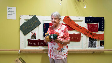 Bildet viser demenssyke Ragnhild på Fosshagen dagsenter i Lier. Hun står foran en tavle med lapper hun har strikket. På lappene står blant annet "alene", "savn" og "ærlig".