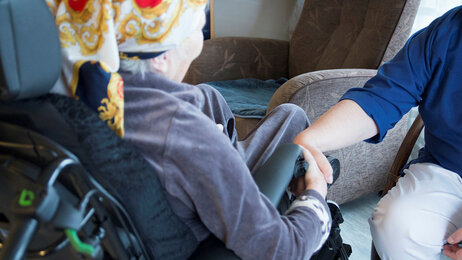 En gamel dame som sitter i en stol og holder en pleier i hånden.