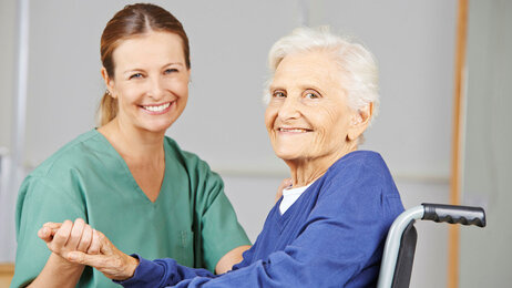 Bildet viser en sykepleier som holder en eldre pasient i hånda. Pasienten sitter i rullestol, og begge smiler.
