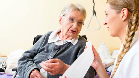 Bildet viser en eldre dame hjemme hos seg selv, og en sykepleier som ruller ut en bandasje mens hun snakker med pasienten. 