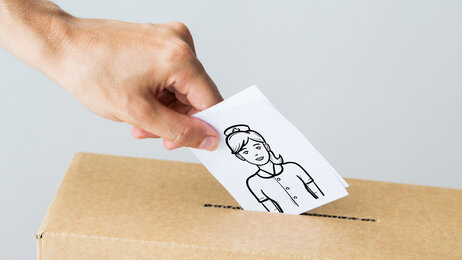 Bildet viser en hånd som slipper en stemmeseddel ned i en papireske. På stemmeseddelen er det bilde av en sykepleier.