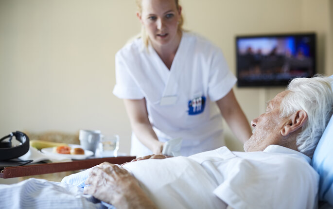 The photo shows a nurse bending over a bed where an older man lies.