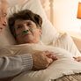 Bildet viser en syk eldre dame som ligger i senga med neseslange. En eldre mann holder henne i hånda og kikker på henne
