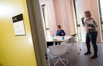 Bildet viser døråpningen til et møterom, der det er to personer. Den ene sitter ved bordet, og den andre står ved siden av og drikker en kopp kaffe.