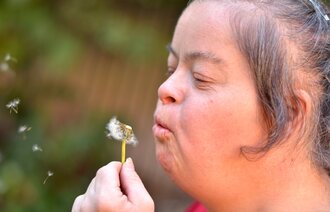 Bildet viser en voksen kvinne med psykisk utviklingshemning som blåser på en løvetann.