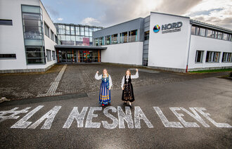 Bildet viser to bunadskledte mennesker utenfor høgskolen i Nesna, på bakken er det skrevet "La Nesna leve".