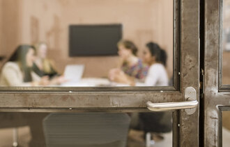 Bildet viser en lukket, gjennomsiktig dør inn til et rom hvor fire personer sitter rundt et bord.