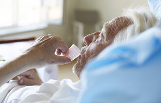 Bildet viser en eldre, syk mann i senga som drikker medisin fra et plastglass