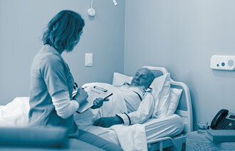 The photo shows a nurse sitting by the bed of an elderly man. She is holding a form in her hand.