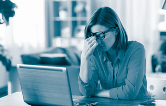The photo shows a woman sitting at home with a laptop in front of her. She looks tired and is wiping her eyes.