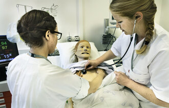 Bildet viser to sykepleiere som står ved en seng. I senga ligger en simuleringsdukke. Den ene sykepleieren lytter etter hjerteslag på dukken