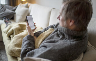 Bildet viser en mann som ligger under et pledd på sofaen og ser på mobilen
