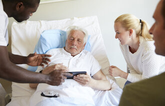 Bildet viser en syk, eldre mann i sengen. En sykepleier står over ham med et nettbrett. En ung dame er pårørende ved sengekanten