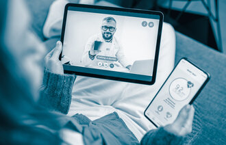 The photo shows a woman sitting in a sofa talking to a doctor via her Ipad. In the other hand she is holding a mobile phone where she is sending her vital data via an app