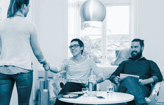 The photo shows a young woman standing with her back towards the reader. She is shaking hands with a smiling woman sitting by a small table. Beside her there is a smiling man. There are books and folders in the bookcase up against the wall..