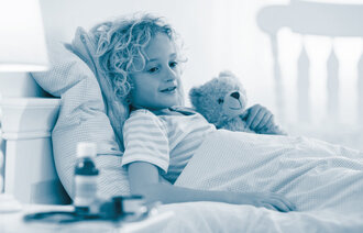 The photo shows an ill, little boy lying in his bed. In his handen he is holding a teddy bear. There are medicine bottles on the bedside table.