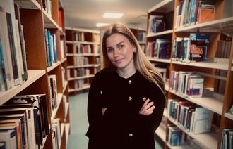 Bildet viser Cathrine Emilie Valgermoe Forren. Hun står i et bibliotek, ser mot kamera og har armene i kors.