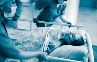 The photo shows a patient on a stretcher carried by running health professionals. The patient is wearing an oxygen mask.