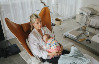 The photo shows a young mother sitting in a chair in her living room. She looks tired and holds her newborn baby in her lap.