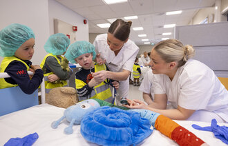 Bildet viser en gruppe med barn som står sammen med to sykepleierstudenter. De undersøker en kose-orm med et stetoskop.