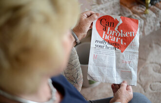 Bildet viser en eldre kvinne som holder opp et avisutklipp hvor det står "can a broken heart really harm your heart?"