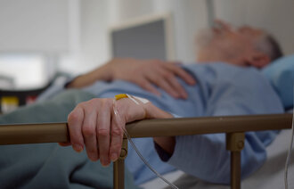 The photo shows an older man lying in a hospital bed. His left hand is resting on the bed rail. In his hand is a venous access port.