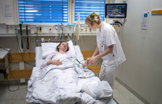 Bildet viser en sykepleier som står ved sengen til en pasient på sykehus