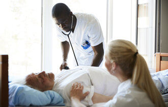 The photo shows an older patient lying in a hospital bed. A male nurse is bending towards him listening with a stethoscope. A female nurse is sitting beside the bed.