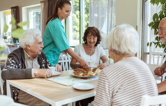Bildet viser fire eldre som sitter rundt et bord. En sykepleier serverer rundstykker til dem