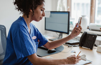 The photo shows a nurse having a video chat on her mobile phone. She is sitting at her desk writing a note.