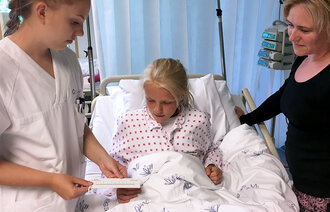 Bildet viser en sykepleier som står ved siden av sykehussengen. I den ligger ei jente. Sykepleieren viser jenta en smerteskala. Moren står til høyre for sengen.