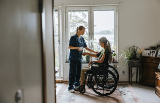 The phowo shows a nurse standing in front of a woman in a wheel chair. The nurse is monitoring her blood pressure. The are in front of a window in the patient's living room