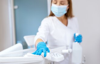 The photo shows a nurse wearing a mask and gloves. She is holding a spray can in her hand, wiping with a cloth.