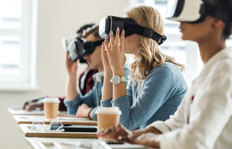 The image shows students with VR glasses. They are sitting by their desks in a class room