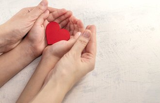 The image shows a pair of female hands gently holding a child’s hands. The child is holding a heart in their hands