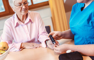 The image shows a nurse monitoring the blood sugar of an older woman in her home