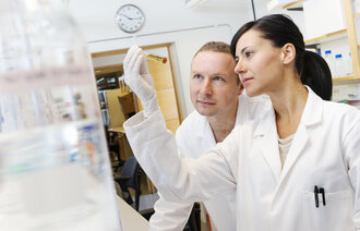 The image shows a woman and a man in white laboratory coats holding a test tube with urine up to the light