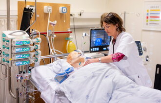 The image depicts a simulation mannequin connected to a ventilator. A nurse is leaning over the bed.