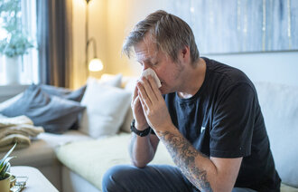 Bildet viser en mann som sitter i en sofa og snyter seg.