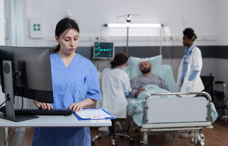 Bildet viser en sykepleier som står på et pasientrom foran en PC. Foran henne ligger et skjema. Bak henne ligger en pasient i senga med to sykepleiere ved sengekanten