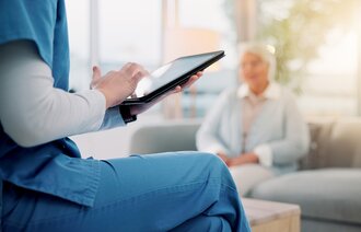 Bildet viser en sykepleier som sitter med en iPad med en pasient i bakgrunnen