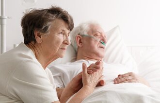 Bildet viser en kone som sitter ved sykesengen til sin døende mann