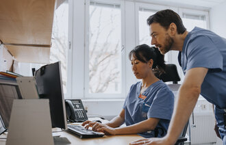 Bildet viser en kvinnelig sykepleier som sitter foran en PC. En mannlig sykepleier står bak henne og kikker på skjermen