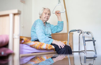 Gammel dame på pleiehjem sitter på sengen med rullator foran. Rutete pledd. 
