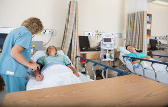 En sykepleier undersøker en pasient på sengepost. En annen pasient ligger i sengen ved siden av.