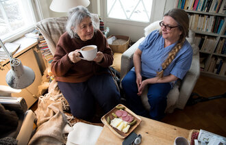 Bildet viser en beboer som drikker kaffe sammen med hjemmesykepleieren. Foran beboeren står et fat med påsmurte brødskiver.