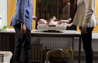 Bildet viser en mor som er på helsestasjonen, der babyen veies av helsesøster.